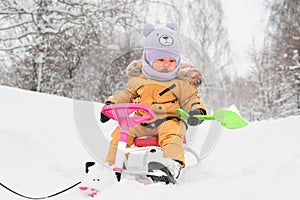 Toddler 12-17 months old with a green toy shovel in his hands sits on a children`s snowcat in a winter park