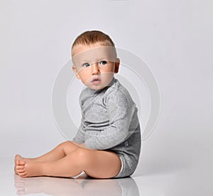 Toddler one-year-old baby boy in diaper and grey one-piece bodysuit with long sleeves sits side and looking to camera