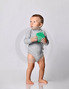 Toddler one-year-old baby boy in diaper and grey one-piece bodysuit with long sleeves stands holding green toy block