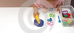 Toddler sculpts from colored plasticine on a white table. The hand of a small child squeezes pieces of colored