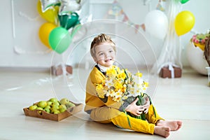 Toddler sits on the floor in a yellow suit with bare feet on the background of a decorated festive room