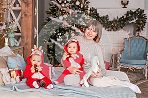 Toddler twins in red reindeer santa claus costumes are sitting at home with their mother against background of christmas tree