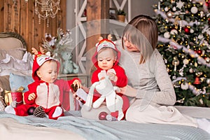 Toddler twins in red reindeer santa claus costumes are sitting at home with their mother against background of christmas tree