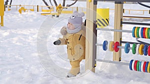 Toddler in yellow warm clothes 12-17 months to learn to walk in the snow on the playground.