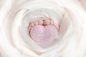 Toes of a newborn baby hug a pink heart toy on a white background