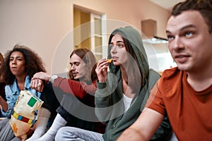 Together. Group of teenagers eating snacks and talking while having fun, relaxing together, sitting on the coach at home