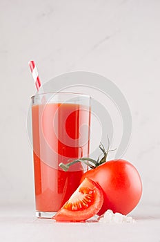 Tomato beverage in glass with branch tomatoes, straw, salt and juicy slice on white wood table, closeup, vertical.