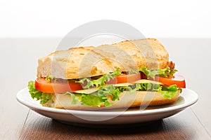 Tomato, cheese and salad sandwich from fresh baguette on white ceramic plate on bright light brown wooden table