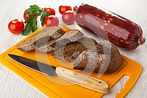 Tomato, parsley, smoked sausage, slices of bread, knife on plastic cutting board on wooden table