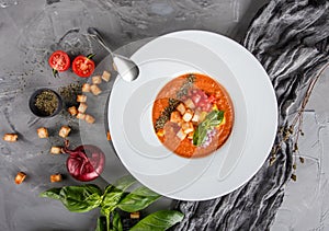 Tomato gazpacho soup with basil, croutons, herbs and vegetables on grey background, Spanish cuisine.