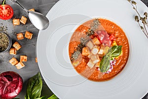 Tomato gazpacho soup with basil, croutons, herbs and vegetables on grey background, Spanish cuisine.