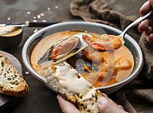 Tomato gazpacho soup with mussels, squid, shrimp in bowl over dark wooden background. Hands holding a spoon with prawn and soup.