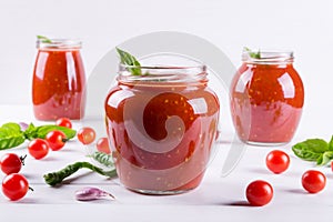 Tomato ketchup sauce with cherry tomatoes and red hot chili peppers, garlic and herbs in a glass jar on white background.