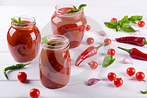 Tomato ketchup sauce with cherry tomatoes and red hot chili peppers, garlic and herbs in a glass jar on white background.