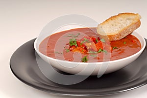 Tomato, red pepper, basil soup in white bowl with bread on a light gray background