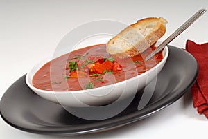 Tomato, red pepper, basil soup in white bowl with bread and spoon on a light gray background