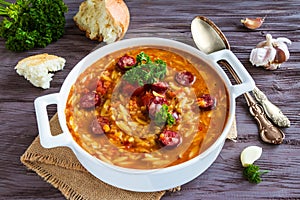 Tomato soup with orzo and smoked sausages in white casserole on wooden rustic table. Fresh bread and parsley, vintage spoon