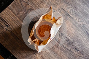 Tomato soup in a paper package on a wooden table. Paper bag. Takeaway food. Plastic cap.