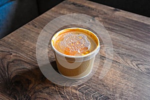 Tomato soup in a paper package on a wooden table. Paper bag. Takeaway food. Plastic cap.