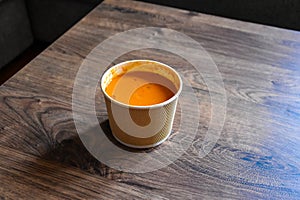 Tomato soup in a paper package on a wooden table. Paper bag. Takeaway food. Plastic cap.