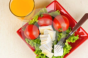 Tomatoes and cheese slices lie on a sheet of fresh salad. A glass of orange juice stands side by side on the table.