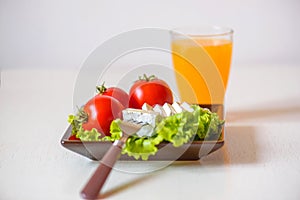 Tomatoes and cheese slices lie on a sheet of fresh salad in a plate. A glass of orange juice stands on the table.