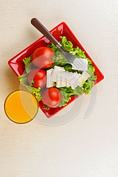 Tomatoes and cheese slices lie on a sheet of fresh salad in a red plate. A glass of orange juice stands side by side on the table.