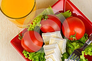 Tomatoes and cheese slices lie on a sheet of fresh salad in a red plate. A glass of orange juice stands side by side.