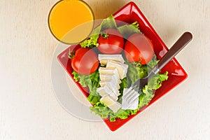 Tomatoes and cheese slices lie on a sheet of fresh salad in a red plate. A glass of orange juice stands side by side.