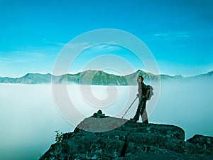 Toned image of an adult woman standing on top of a mountain with a backpack and Alpenstocks against mountains in a fog