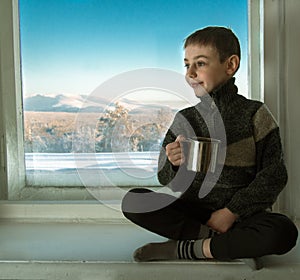 Toned image of a little boy sitting on an old window sill next to the window and holding in his hand a metal cup against the