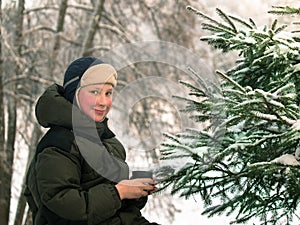 Toned image of a little boy standing next to the fir tree and holding a mug on blurred background trees