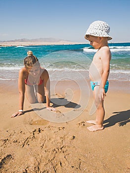 Toned image of young mother playing with her little child boy on wet sand at sea beach. Family relaxing and having good