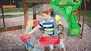 Toned photo of cute little toddler boy playing with toy truck and trailer on playground at park