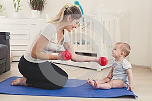 Toned photo of cute smiling baby boy looking at mother sitting on fitness mat and practicing fitness