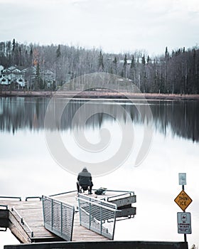 Toned photo fisherman sitting on small backpacking chair on fishing dock with barrier edge protection, autumn forest with row of