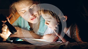 Toned portrait of young mother holding flashlight and reading bedtime story book to her little son lying under blanket