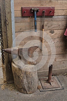 Tools and chains hanging on pegs on a rough wooden wall made of planks next to an anvil in a barn.