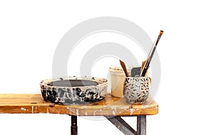 Tools for pottery with potters wheel are standing on the wooden table in messy studio, white isolated