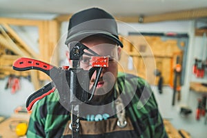 Tools for wood workshop. Male carpenter in black cap blinking at camera and holding bar clamp in front of his face.