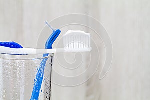 Toothpaste on toothbrush and interdental brush on wet glass on blurred wooden background in bathroom