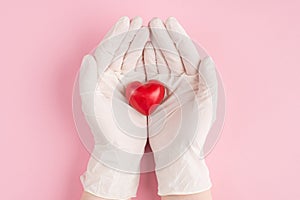 Top above overhead pov first person close up view photo of red heart in female doctor`s hands in gloves isolated on pastel pink