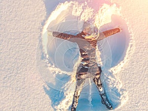 Top aerial view of young happy smiling girl making by arms snow angel figure and lying in snow, winter outdoor activity