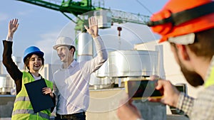 On the top of construction site charismatic guy take pictures for two young engineers lady and man day posing in front