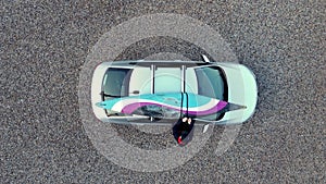 Top down aerial drone shot as man loads a slalom kayak onto roof rack bars on a silver car and tightens the loading straps