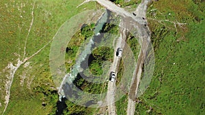 A top-down aerial view of two cars driving along a dirt road along a mountain river to the bridge.