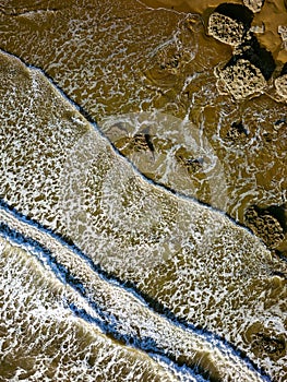 Top down aerial view of waves breaking onto a sandy beach with large boulders Southerndown