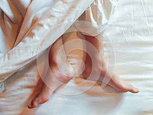 The top-down of both feet of a person on a white bed with a white blanket covering the upper leg