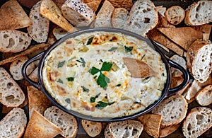 Top down close up of a hot artichoke dip garnished with Italian parsley surrounded by bread and pita pieces for dipping.