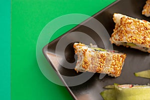 Top down close up view of sushi rolls with sesame seeds and cucumber on a black square plate. Flat on a bright green and light